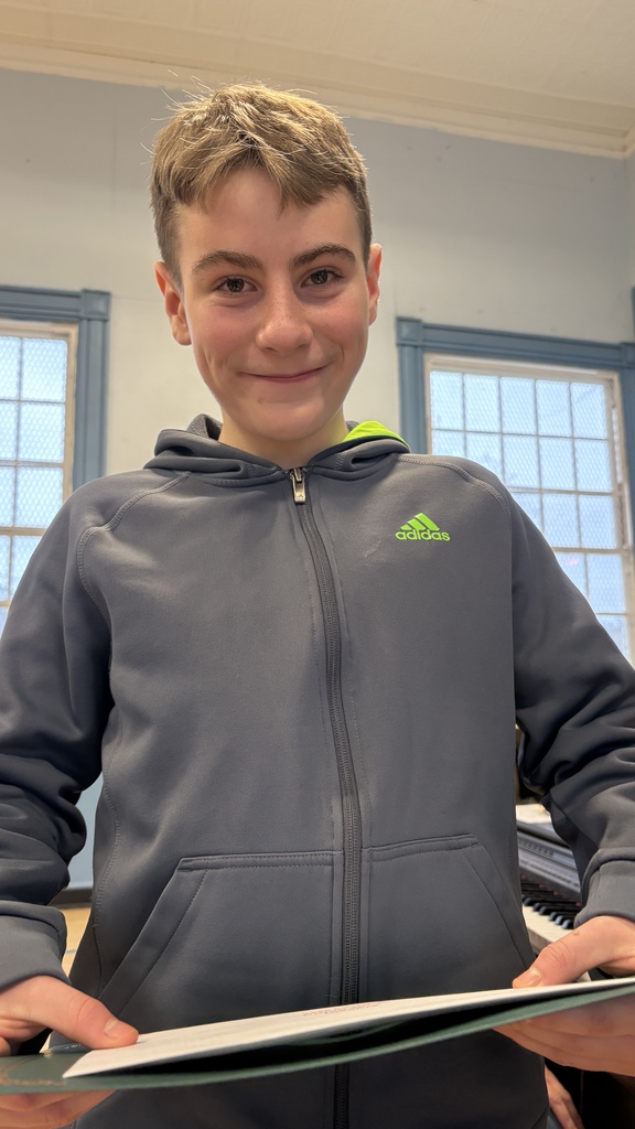 Student in a gray Adidas zip-up jacket smiles while holding a certificate folder in the school gym during the recognition ceremony.
