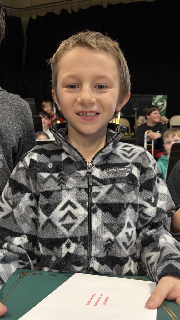 Smiling student wearing a gray patterned Columbia fleece stands in the gym holding a green folder and Student of the Month envelope.