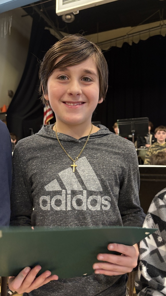 Student in a dark gray Adidas hoodie and gold cross necklace smiles while holding a green folder during a school recognition event.