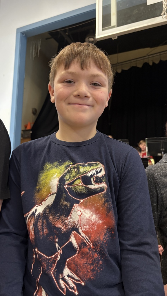 Student wearing a navy shirt with a dinosaur graphic smiles at the camera while holding a green folder at the front of the gym.