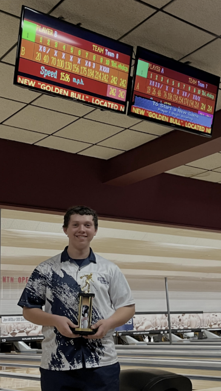 High school student bowler stands in a bowling alley holding a trophy and smiling, with electronic scoreboards and lanes visible in the background.