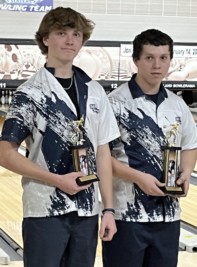 Two high school student bowlers stand side by side at a bowling alley, each holding a trophy and wearing matching team jerseys, with bowling lanes behind them.
