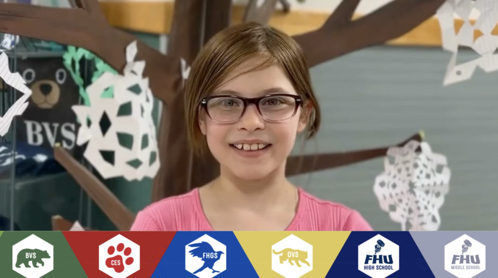 Smiling elementary-age student wearing glasses and a pink shirt stands in front of a decorative tree display with paper snowflakes. A banner along the bottom shows Slate Valley school logos including BVS, CES, FHGS, OVS, FHU High School, and FHU Middle School.