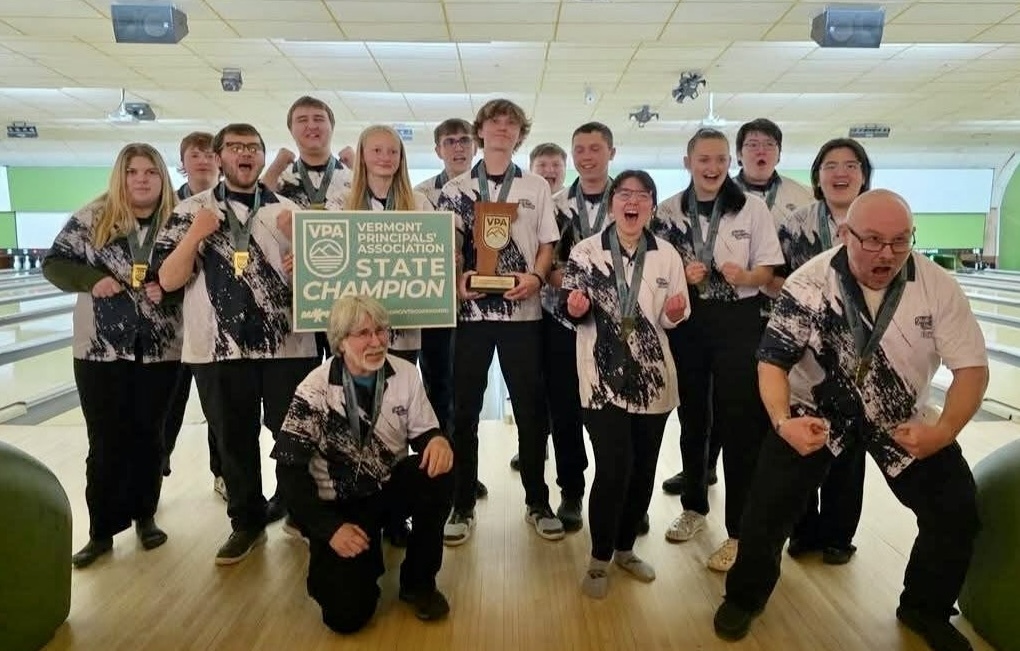A large group of Fair Haven Slater bowling team members celebrate together at a bowling alley, wearing matching team jerseys and medals while holding a Vermont Principals’ Association State Champion sign and trophy.