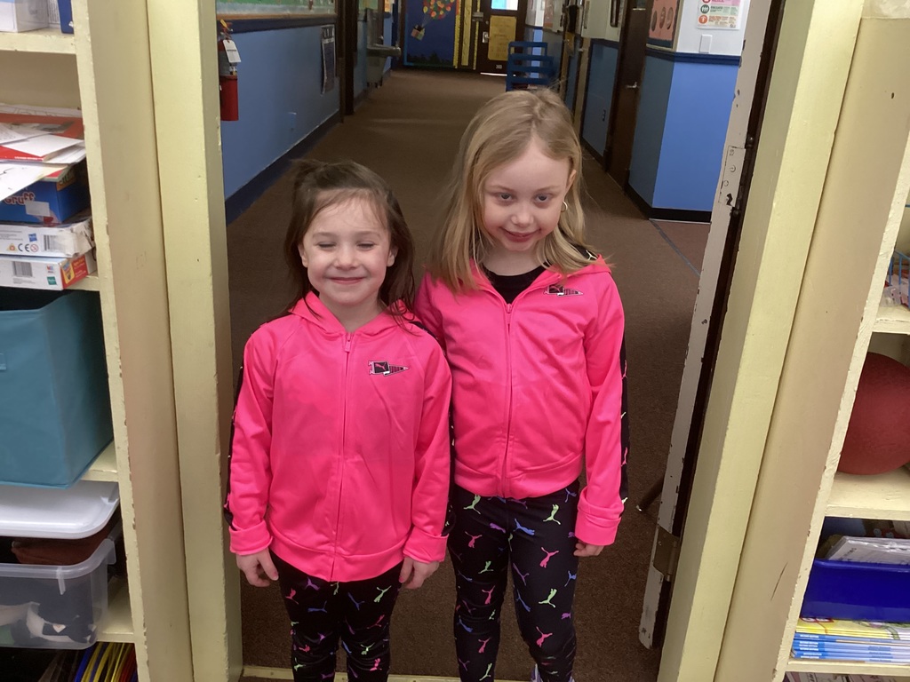 Two kindergarten girls pose together in a hallway wearing matching bright pink zip-up jackets and patterned leggings for Twin Day.