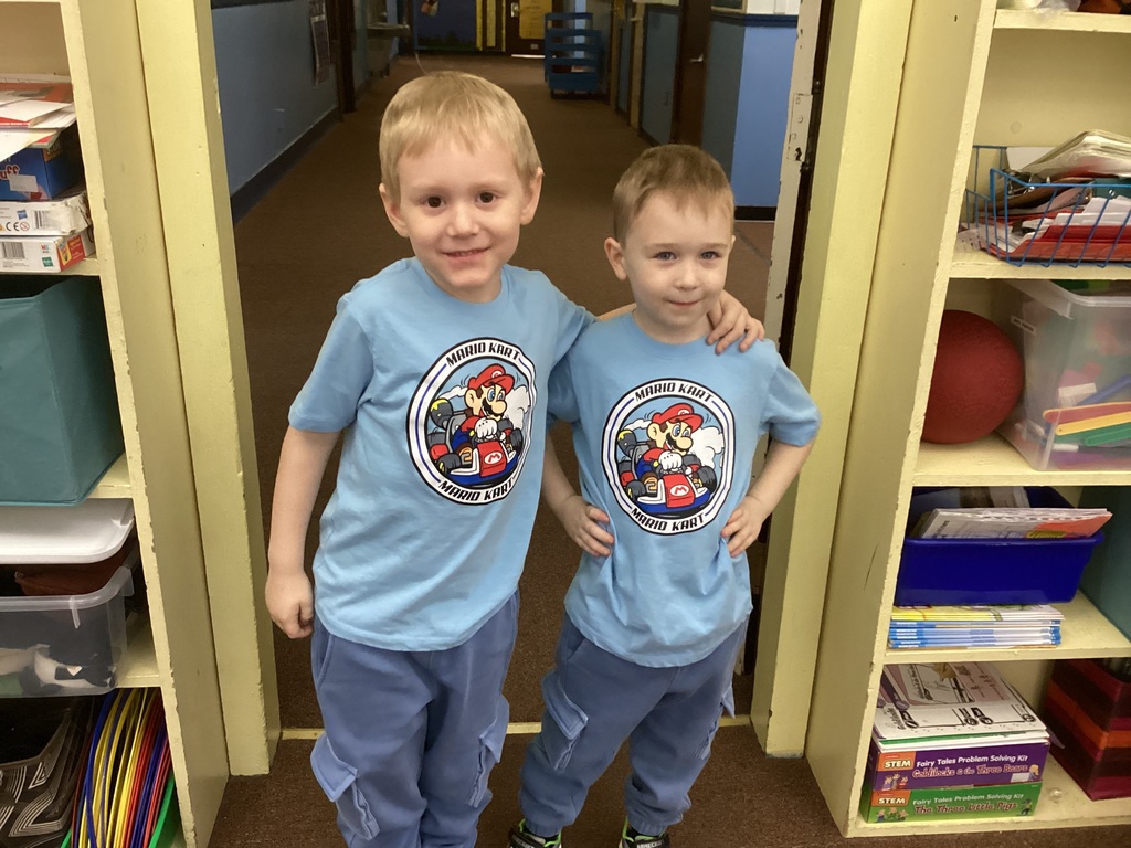 Two kindergarten boys stand side by side in a school hallway wearing matching light blue Mario Kart T-shirts and similar pants.