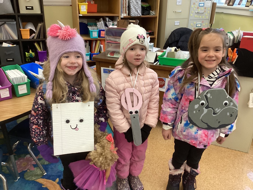 Three kindergarten girls stand in a classroom wearing winter hats and jackets with handmade paper costumes, smiling during Twin Day.