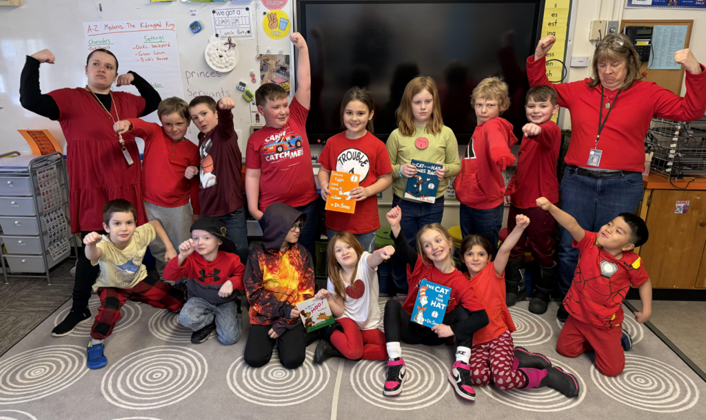 Second grade students and their teacher pose together in a classroom wearing red for Spirit Week and celebrating Dr. Seuss. Several students hold Dr. Seuss books and strike superhero poses while standing and kneeling on a classroom rug.