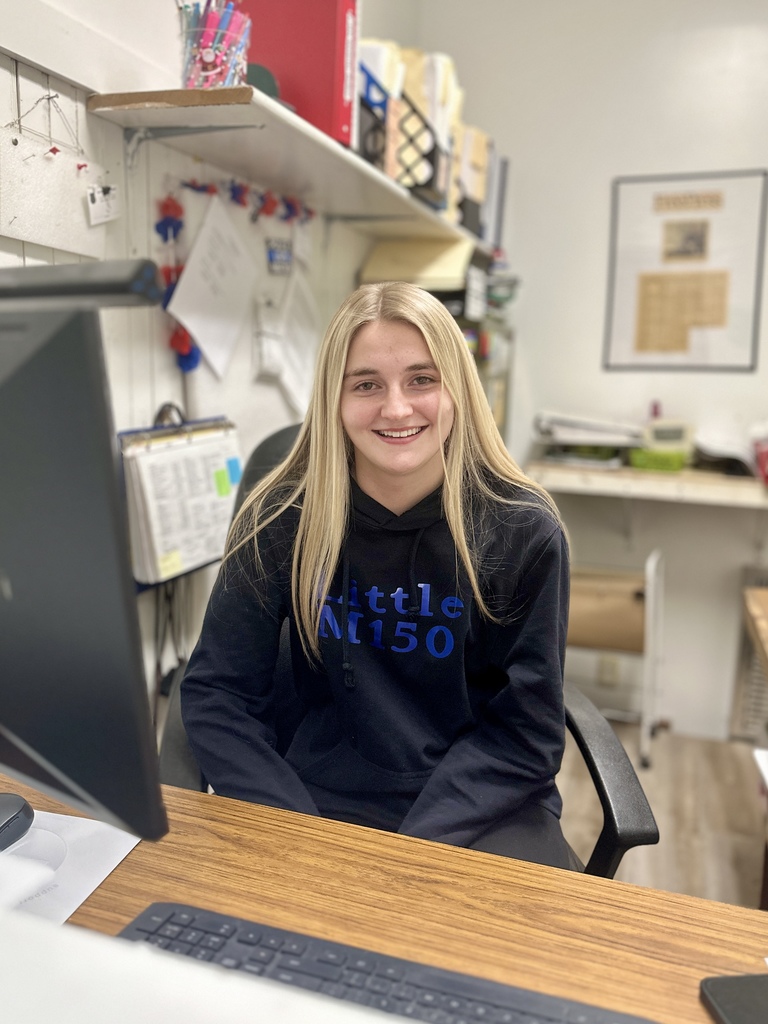 Junior Katie Bouzianis sits at a desk in an office at the Fair Haven Police Department, smiling toward the camera while working at a computer. Office shelves, paperwork, and a bulletin board are visible behind her.
