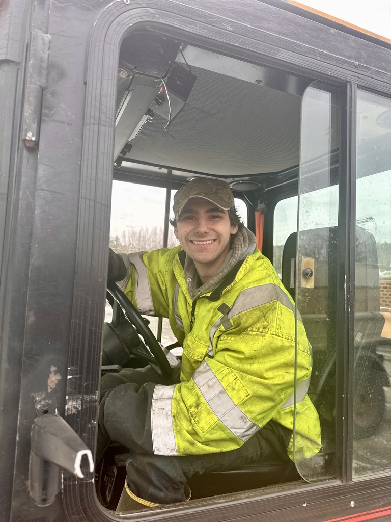 Senior Kaidyn Runnells smiles while seated in the cab of a municipal snowplow, wearing a high-visibility yellow jacket and baseball cap as part of his Work Based Learning placement with the Fair Haven Public Works Department.