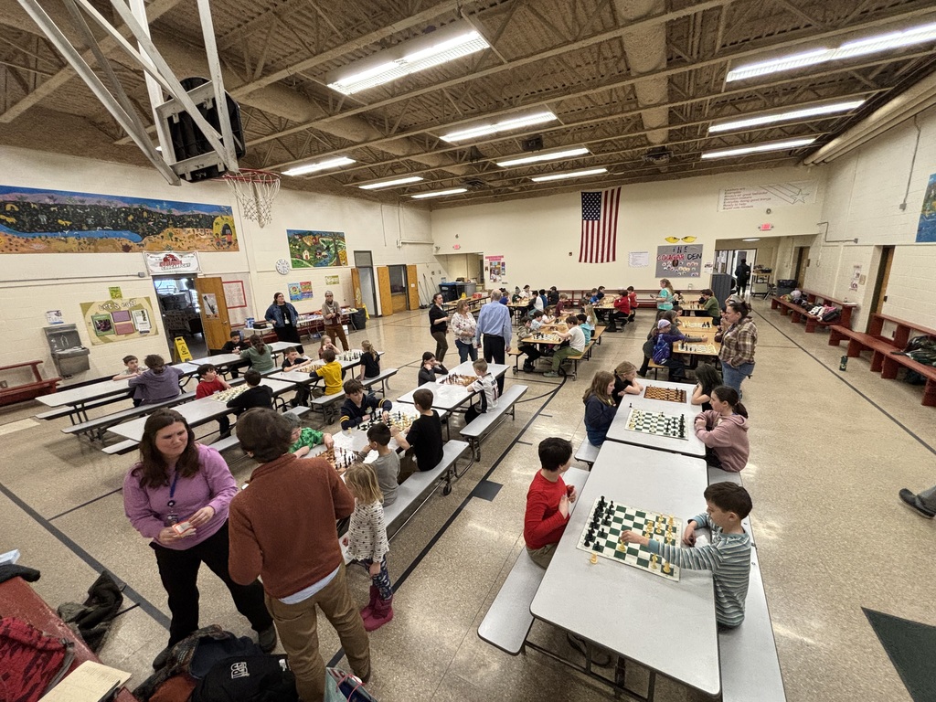 Wide view of a school cafeteria filled with students seated at long tables playing chess. Multiple games are underway while adults and students observe and walk between tables under a basketball hoop and American flag.