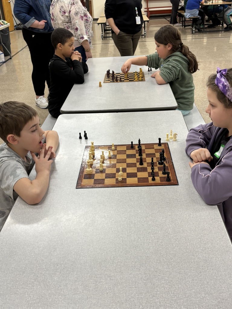 Four elementary-aged students sit around two cafeteria tables playing chess. Two students concentrate on a game in the foreground while another student moves a piece on the board behind them.