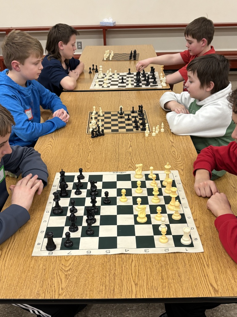 Several students sit around a wooden cafeteria table playing chess in pairs. One student reaches to move a piece while the others watch the board and consider their next moves.