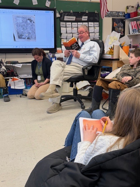 A man reads from a book while students sit around the classroom taking notes or drawing on small papers during a group reading lesson.