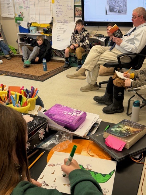 A student sits at a desk drawing on paper with markers while a classroom read-aloud takes place in the background with students seated around the room.