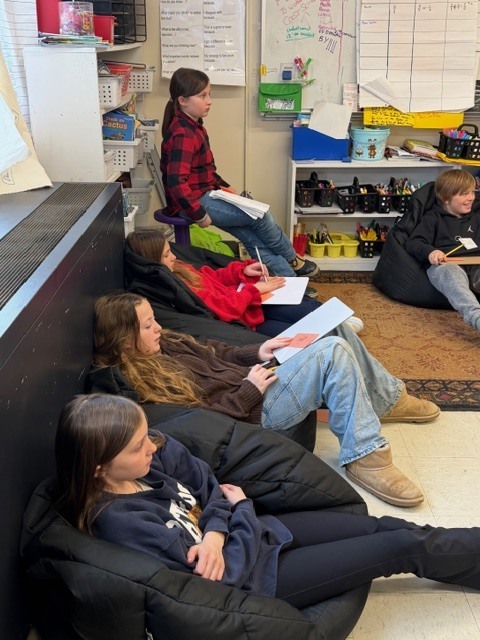 Several students sit in beanbag chairs and on stools along the classroom wall, holding paper and pencils while listening and working during a read-aloud activity.