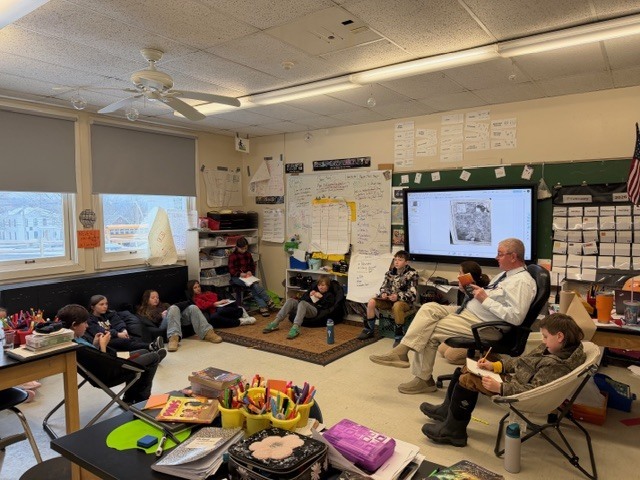 Students sit in a circle around a classroom while an adult reads aloud from a book. Students listen from chairs, beanbags, and the floor, with a digital display and classroom materials visible behind them.
