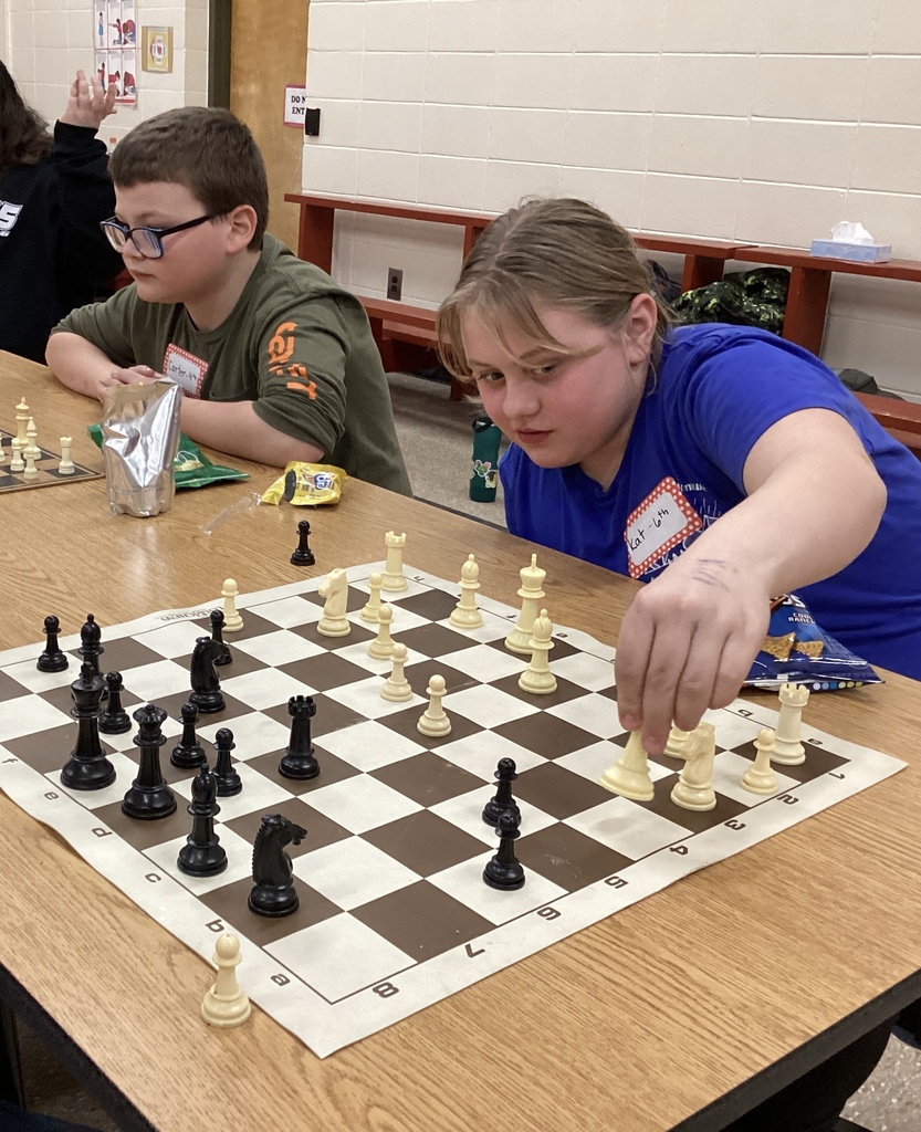 A student carefully moves a white chess piece while concentrating on the board during a chess match at a school chess exhibition.