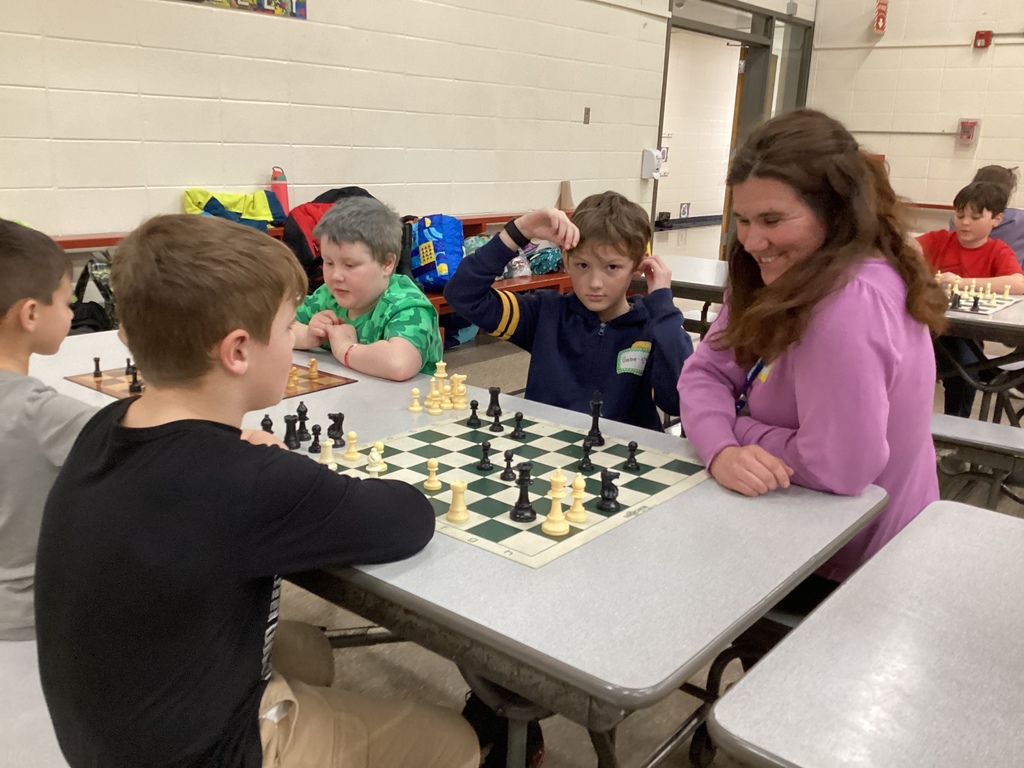 Several students sit at cafeteria tables playing chess matches while an adult volunteer smiles and observes a game.
