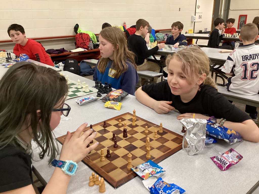 Two students lean over a chessboard in deep concentration while playing a match at a cafeteria table surrounded by other students and snacks.
