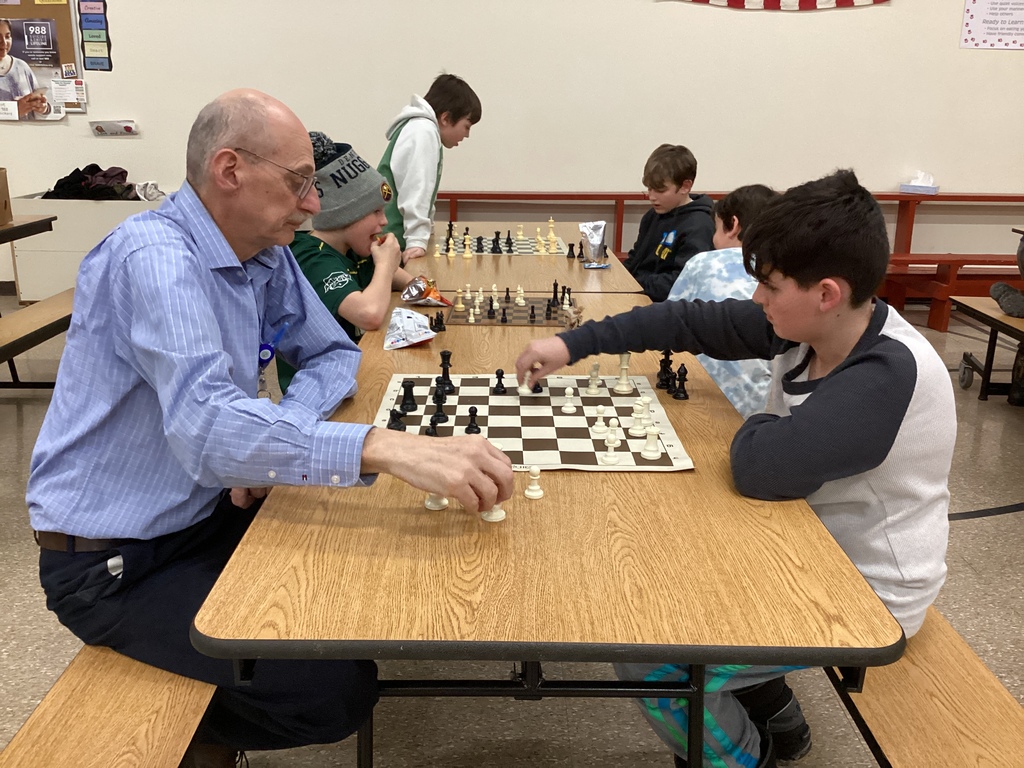An adult volunteer and a student sit across from each other playing chess at a cafeteria table while other students compete in games behind them.