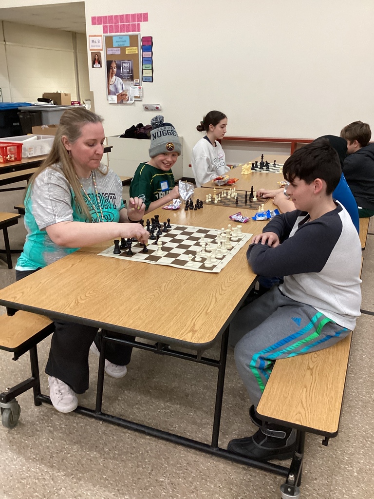 An adult volunteer and a student play chess at a cafeteria table while other students play games in the background during a school chess event.