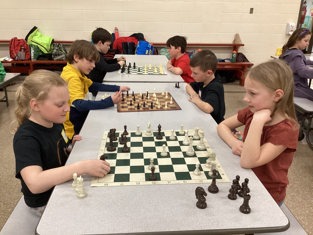 Several students sit at cafeteria tables playing chess matches while an adult volunteer smiles and observes a game.