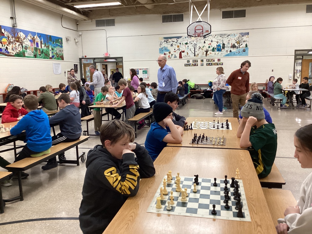 Wide view of the school cafeteria filled with students playing chess games at multiple tables during a chess exhibition event.