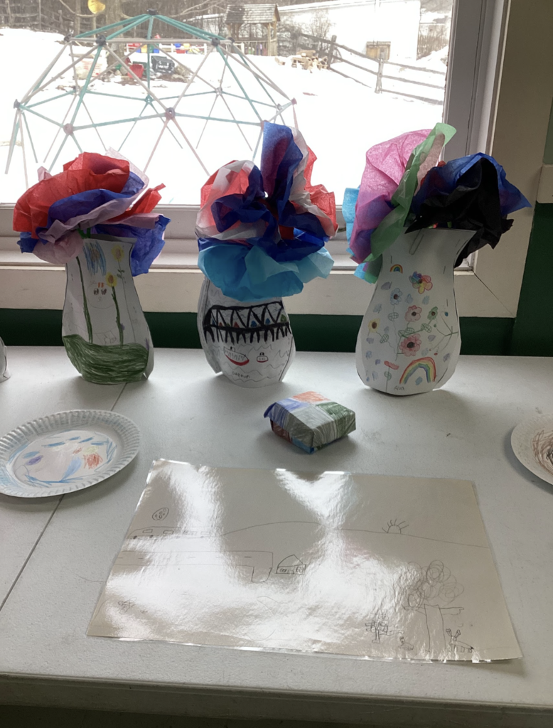 Three student-decorated paper vases filled with bright tissue paper flowers displayed on a table near a window. In front of the vases are student drawings and a small folded paper craft.