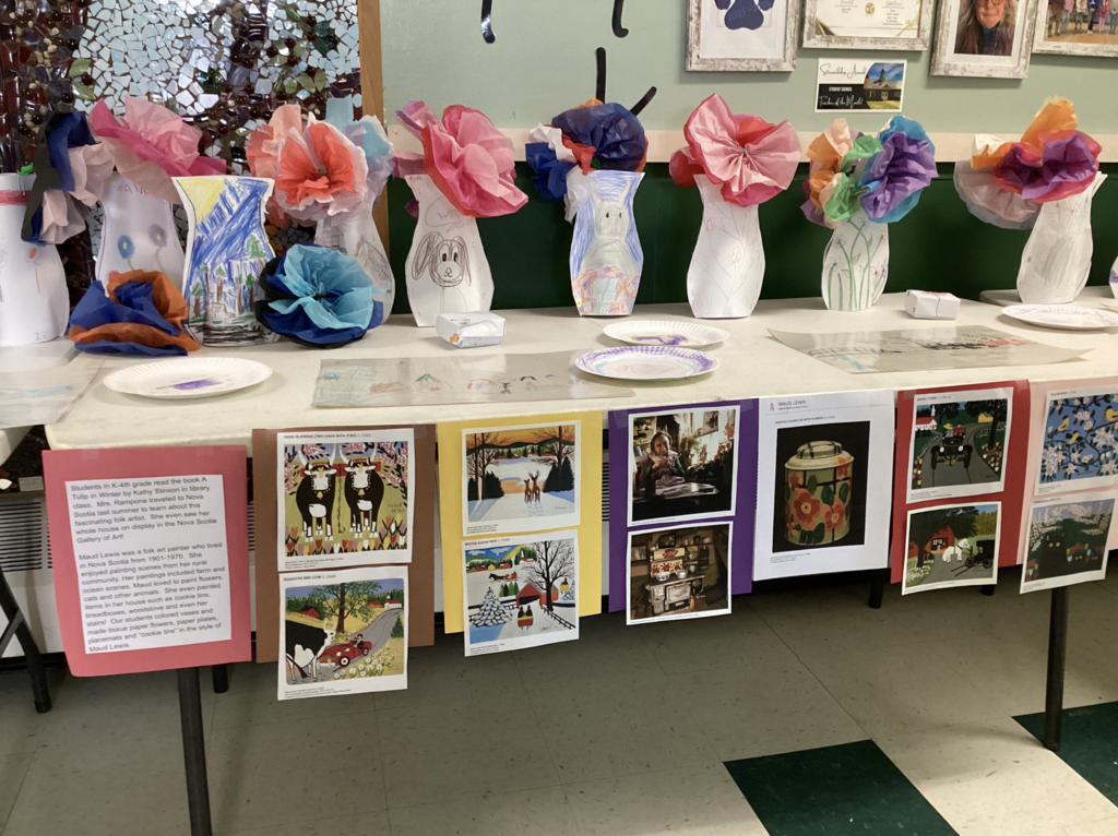 A table display of student art projects featuring decorated paper vases with tissue paper flowers. Beneath the table are posters showing artwork and information about folk artist Maud Lewis.