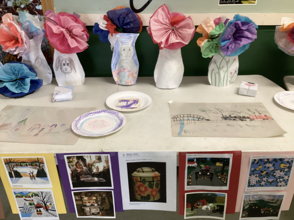 Student art display featuring decorated paper vases with tissue paper flowers arranged across a table. Beneath the display are posters and images highlighting the life and artwork of Maud Lewis.
