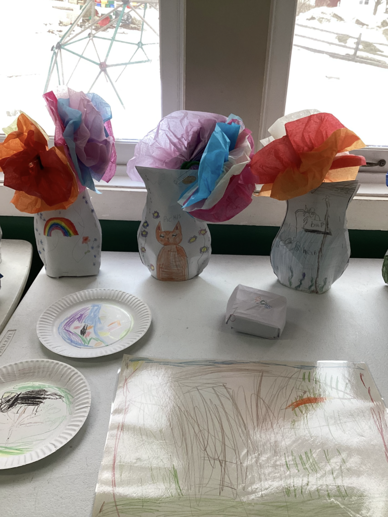 Student artwork displayed on a table by a classroom window, featuring decorated paper vases filled with colorful tissue paper flowers. In front of the vases are drawings, small folded paper crafts, and paper plates with student artwork.