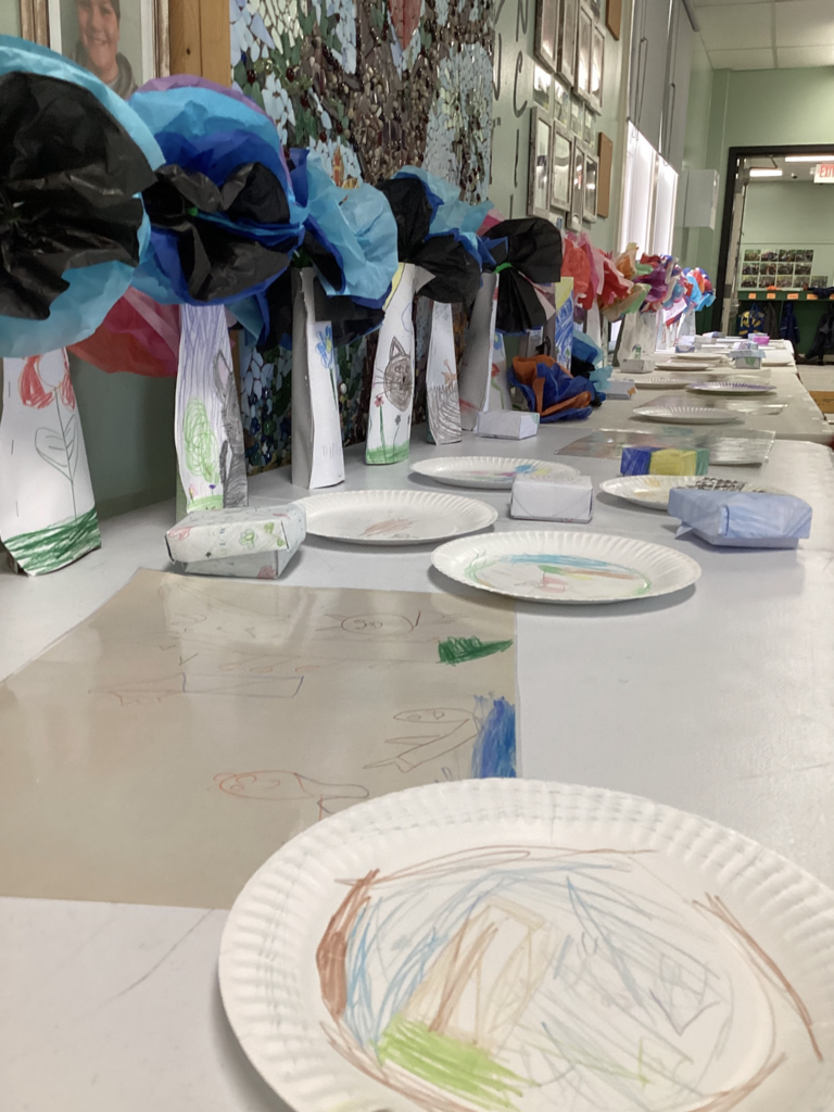 Close-up view of a hallway art display with multiple student-made paper vases topped with colorful tissue paper flowers. Drawings, paper plates with artwork, and small paper crafts are arranged on the table.