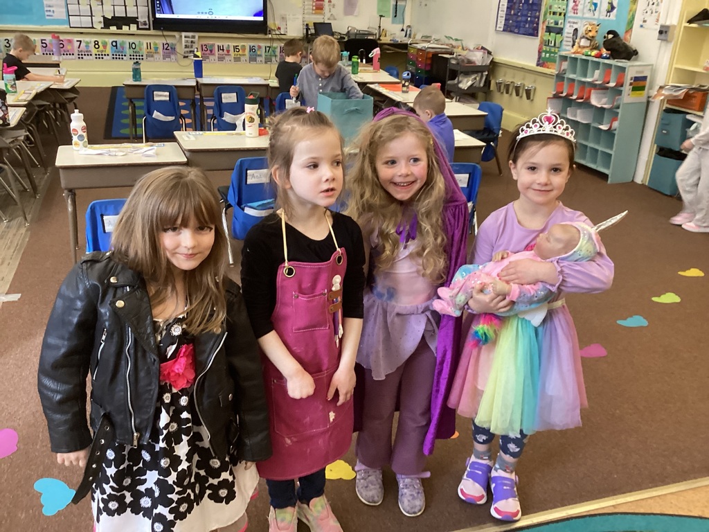 Four kindergarten students stand together in a classroom wearing costumes for Spirit Week. One wears a leather jacket and dress, another an artist apron, another a colorful princess-style outfit, and another a tiara while holding a baby doll.