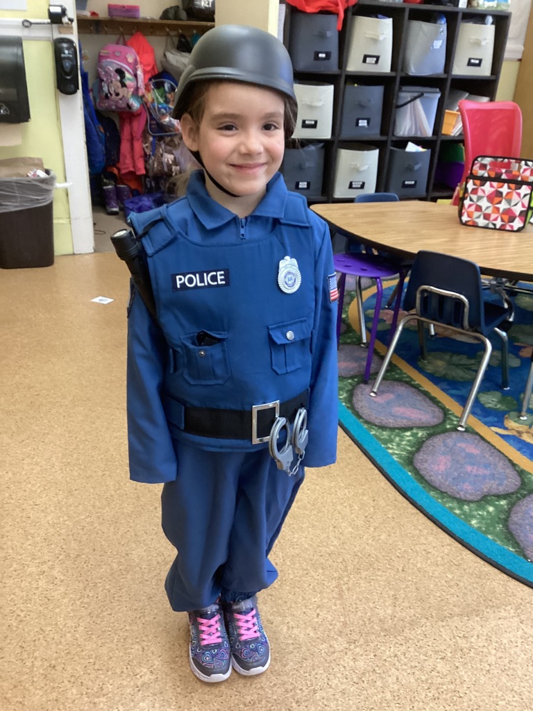 A kindergarten student smiles while wearing a police officer costume, including a helmet, badge, and toy handcuffs, standing in a classroom with cubbies and tables in the background.
