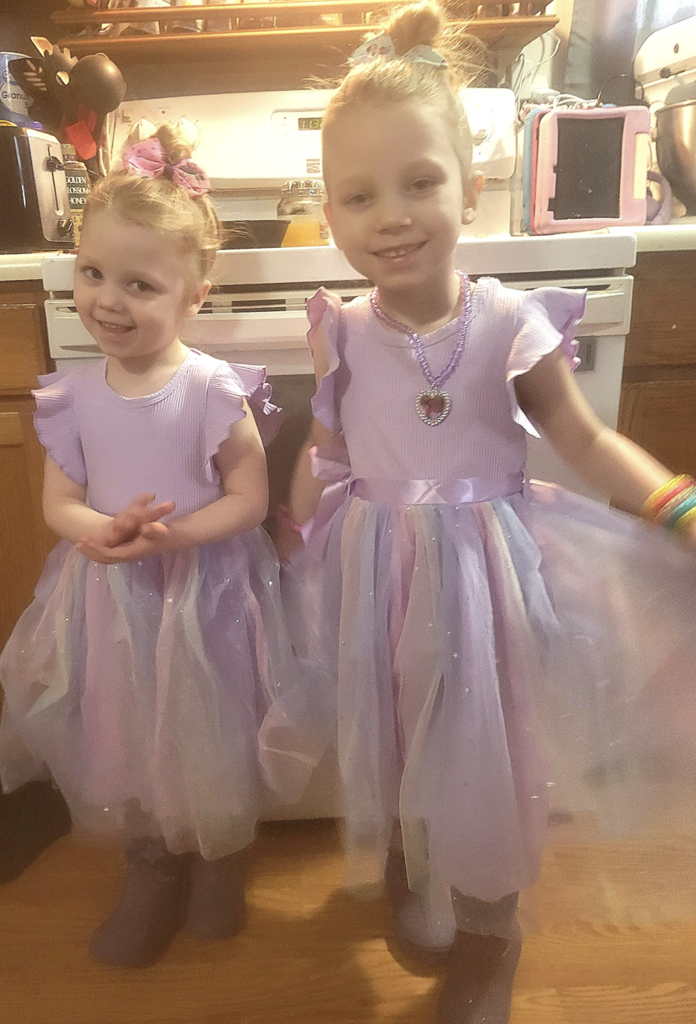 Two young children stand in a kitchen wearing matching pastel purple and pink sparkly dresses with tulle skirts and boots, smiling while posing together.