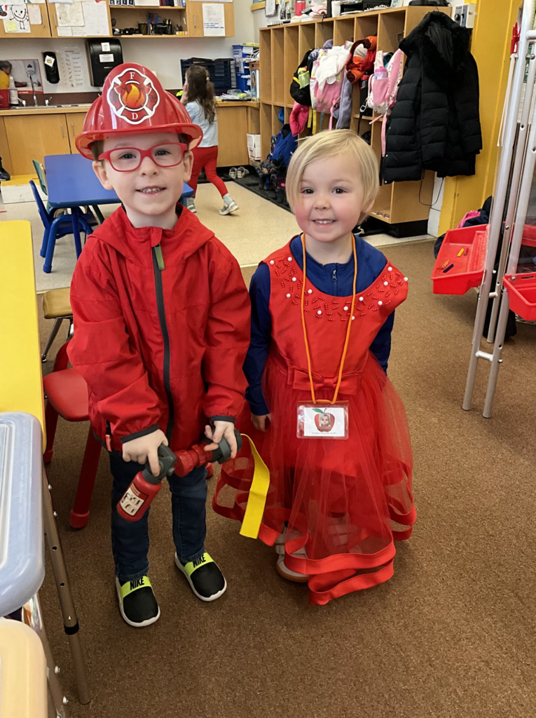 Two preschool students in a classroom dressed for spirit week: one child wearing a red firefighter helmet, red jacket, glasses, and holding a toy fire extinguisher, standing next to a child in a bright red dress with a name badge.