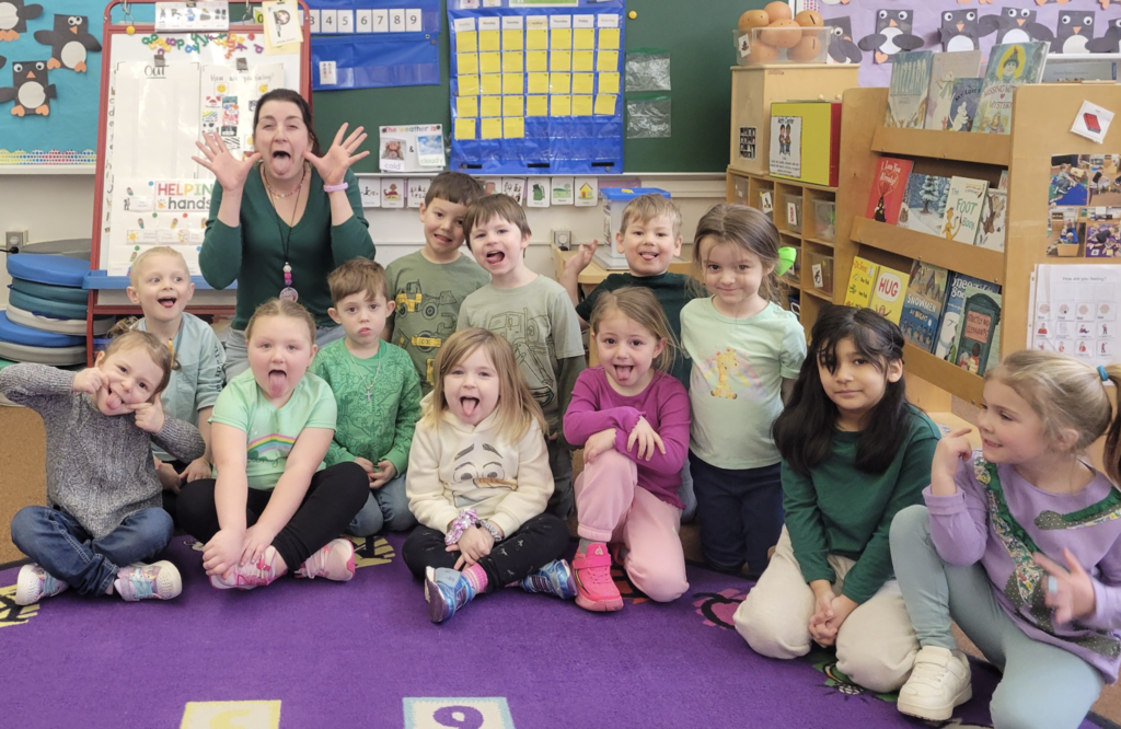 A preschool teacher and group of students sit and kneel on a colorful classroom rug making silly faces and sticking out their tongues while posing for a playful group photo.
