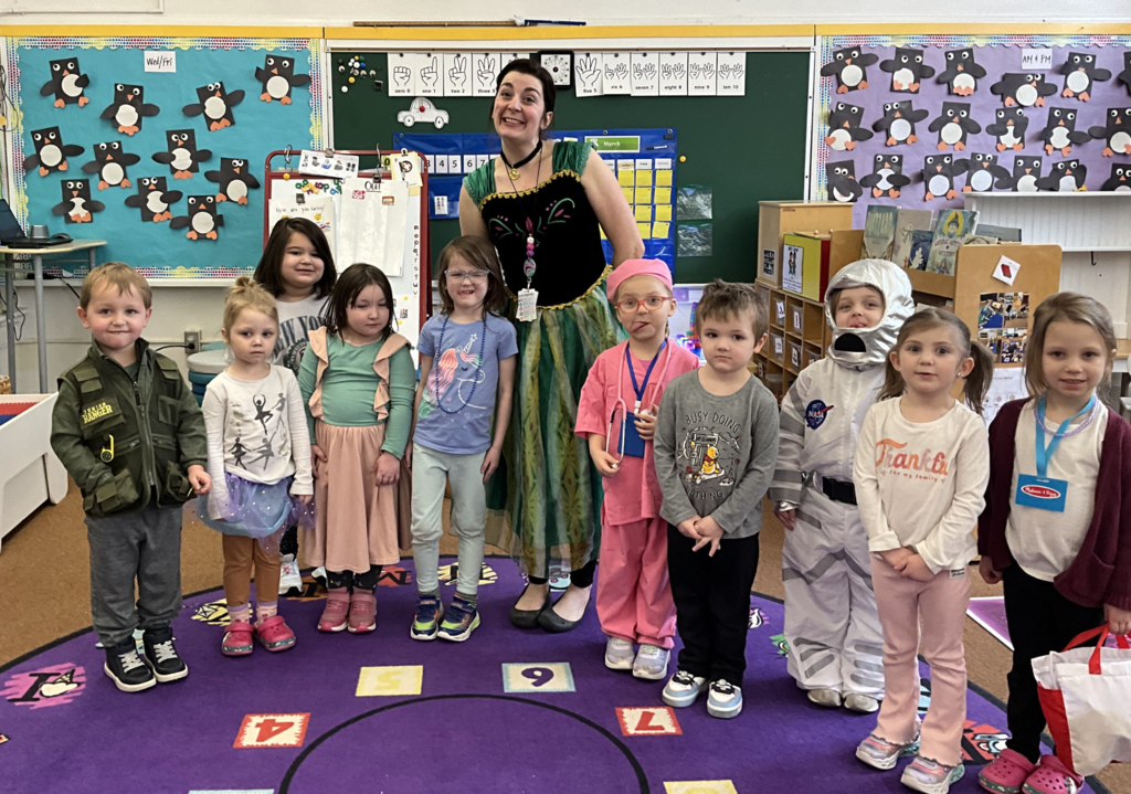 A preschool class stands with their teacher in a classroom while dressed as different future careers, including a doctor, astronaut, dancer, and other costumes. The teacher stands in the middle wearing a green and black dress while the students pose on a colorful classroom rug.