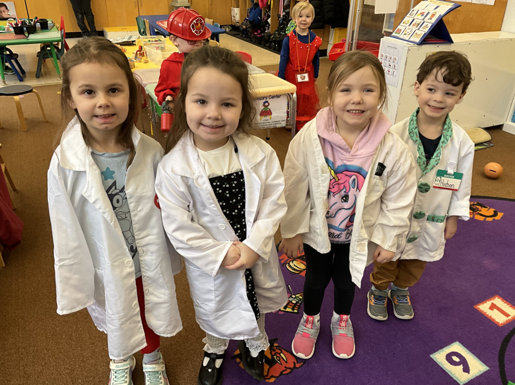 Four preschool students stand together in a classroom wearing white lab coats and medical-themed outfits, pretending to be doctors or scientists. Other students dressed for spirit week can be seen in the background.