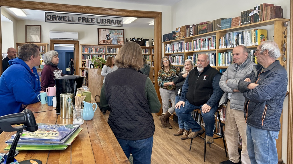 Group of community members gathered inside the Orwell Free Library during a Coffee and Conversation event with Senator Ruth Hardy. Several people stand and sit near bookshelves while listening and participating in discussion. A coffee station with mugs and a coffee maker is visible in the foreground.