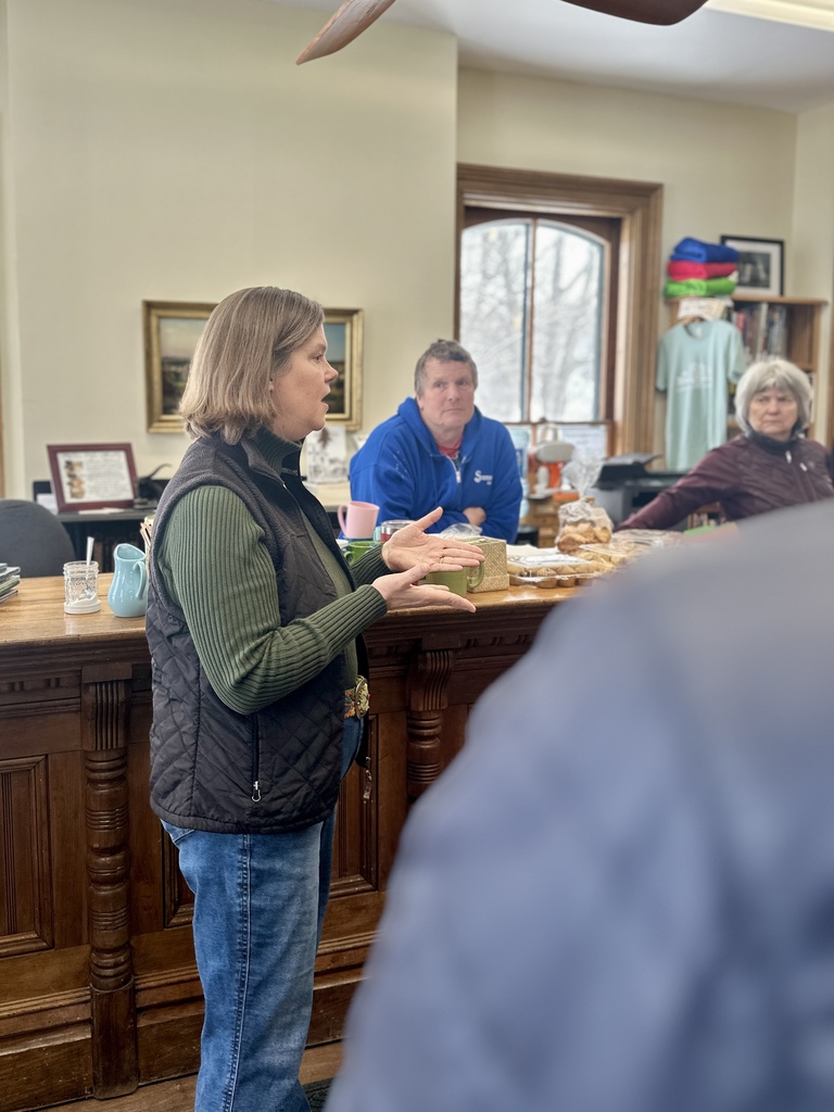 Community members stand inside the Orwell Free Library listening to Senator Ruth Hardy speak during a Coffee and Conversation event. Bookshelves line the walls and people gather with coffee while the senator addresses the group near the front of the room.