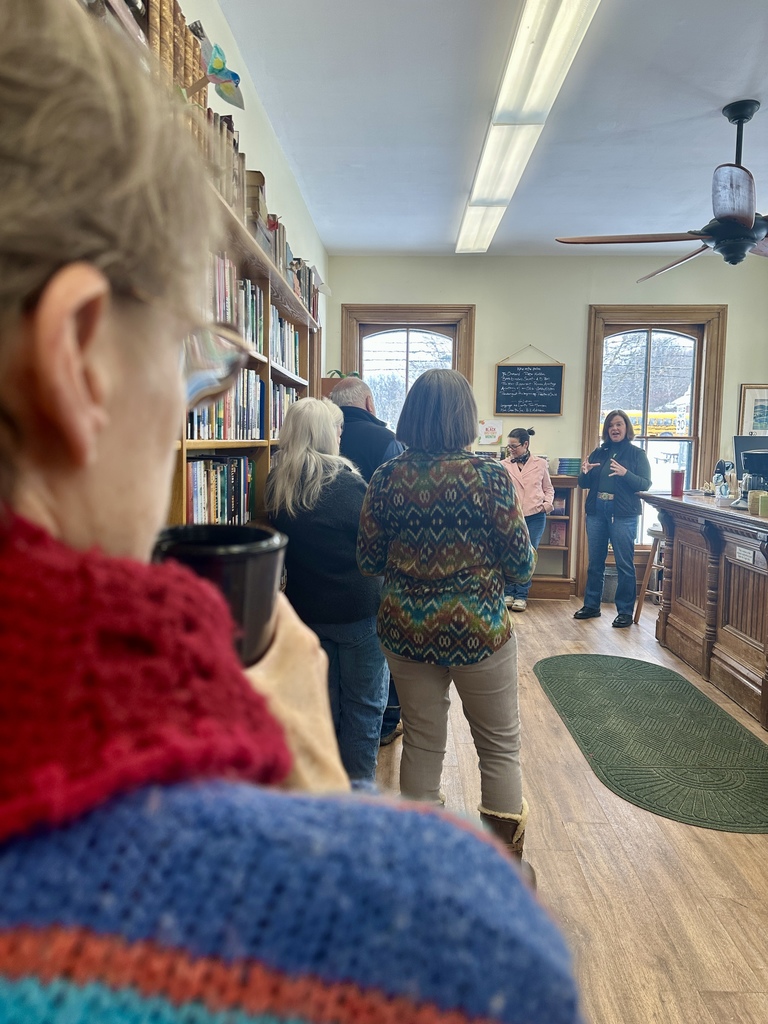 Community members stand inside the Orwell Free Library listening to Senator Ruth Hardy speak during a Coffee and Conversation event. Bookshelves line the walls and people gather with coffee while the senator addresses the group near the front of the room.