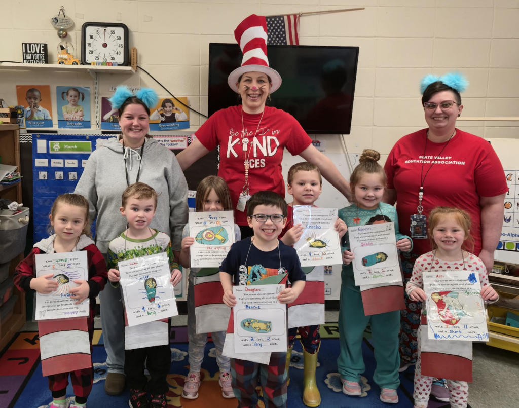 Kindergarten students stand with their teachers dressed as Dr. Seuss characters in a classroom while holding illustrated worksheets from a Dr. Seuss–themed activity.