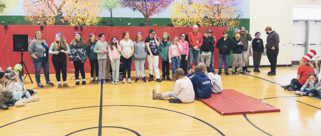 A large group of students wearing colorful glasses stands in a line across the gym during a school assembly while other students sit on the floor watching.