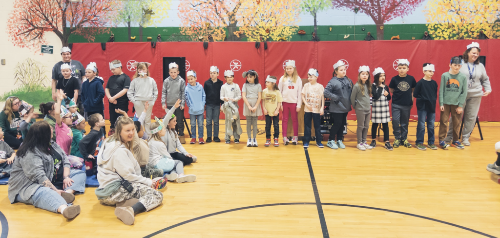 Students stand in a line on the gym floor wearing paper headbands while classmates sit nearby watching during a school assembly activity.
