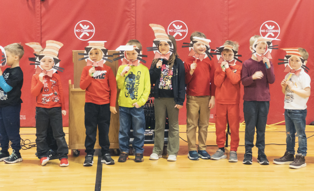 Elementary students stand on a gym floor wearing Cat in the Hat–style paper masks and hats while participating in a Dr. Seuss–themed activity during a school assembly.