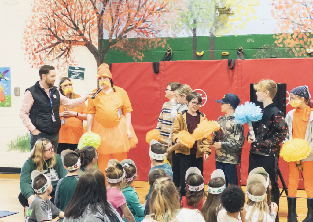 Students and staff dressed in bright orange costumes and holding colorful pom-poms participate in a lively performance during a Dr. Seuss–themed assembly while younger students watch from the gym floor.