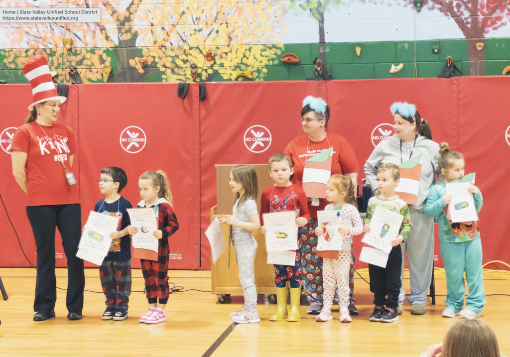 Students stand in a line in a school gym holding papers while staff members dressed as characters from The Cat in the Hat stand nearby during a Dr. Seuss–themed school assembly.