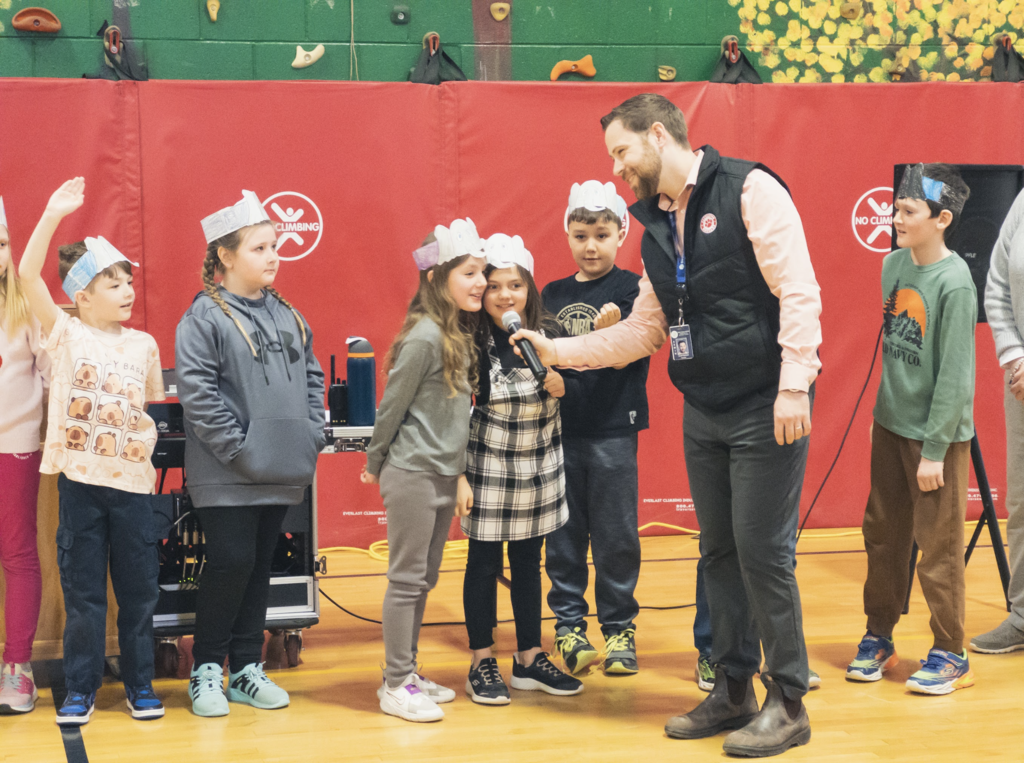 A school staff member holds a microphone for two students wearing paper headbands as they speak during a Dr. Seuss–themed assembly in the gym while other students stand nearby waiting their turn.